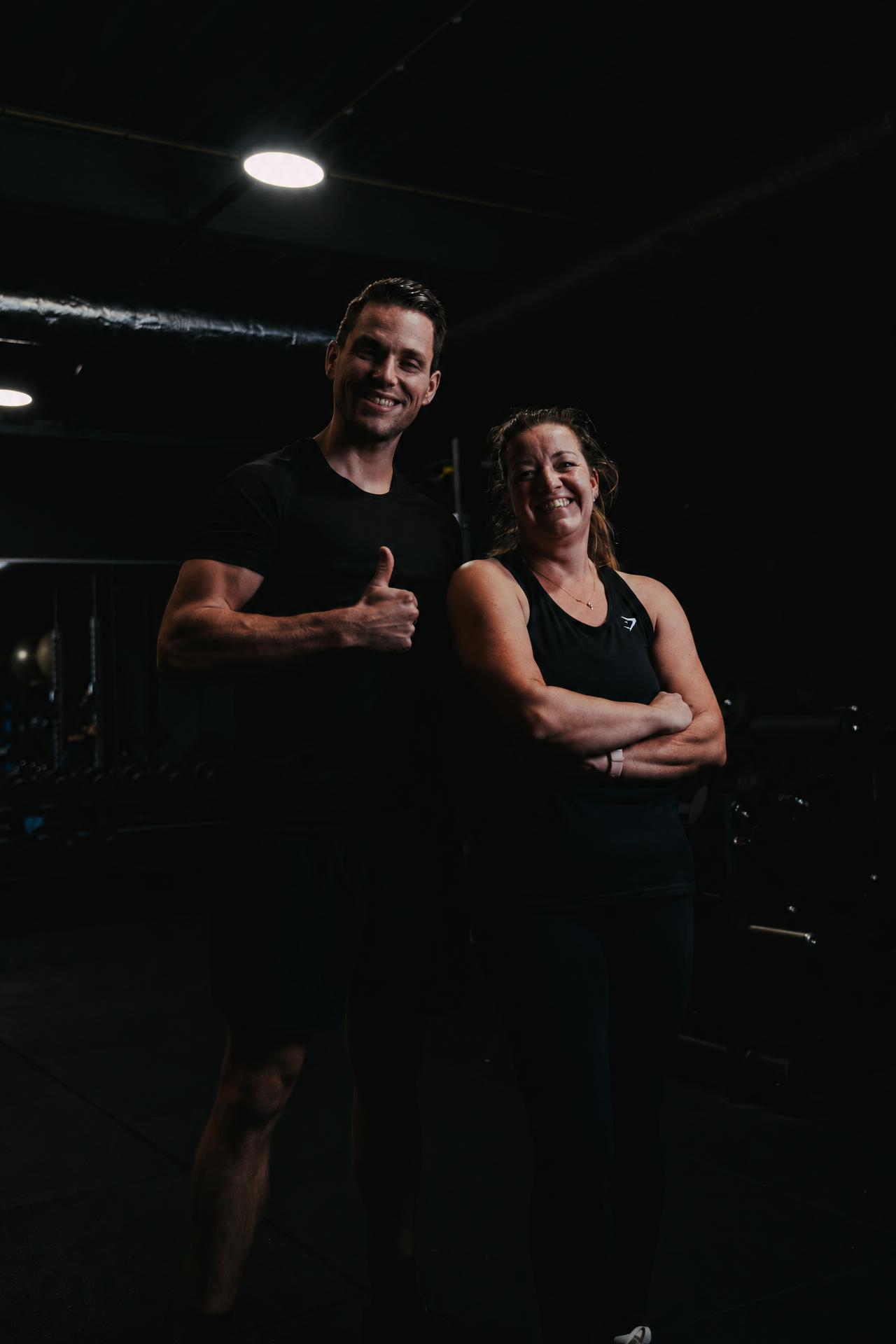 Coach Tijs steekt lachend zijn duim op naast een tevreden cliënt in de gym